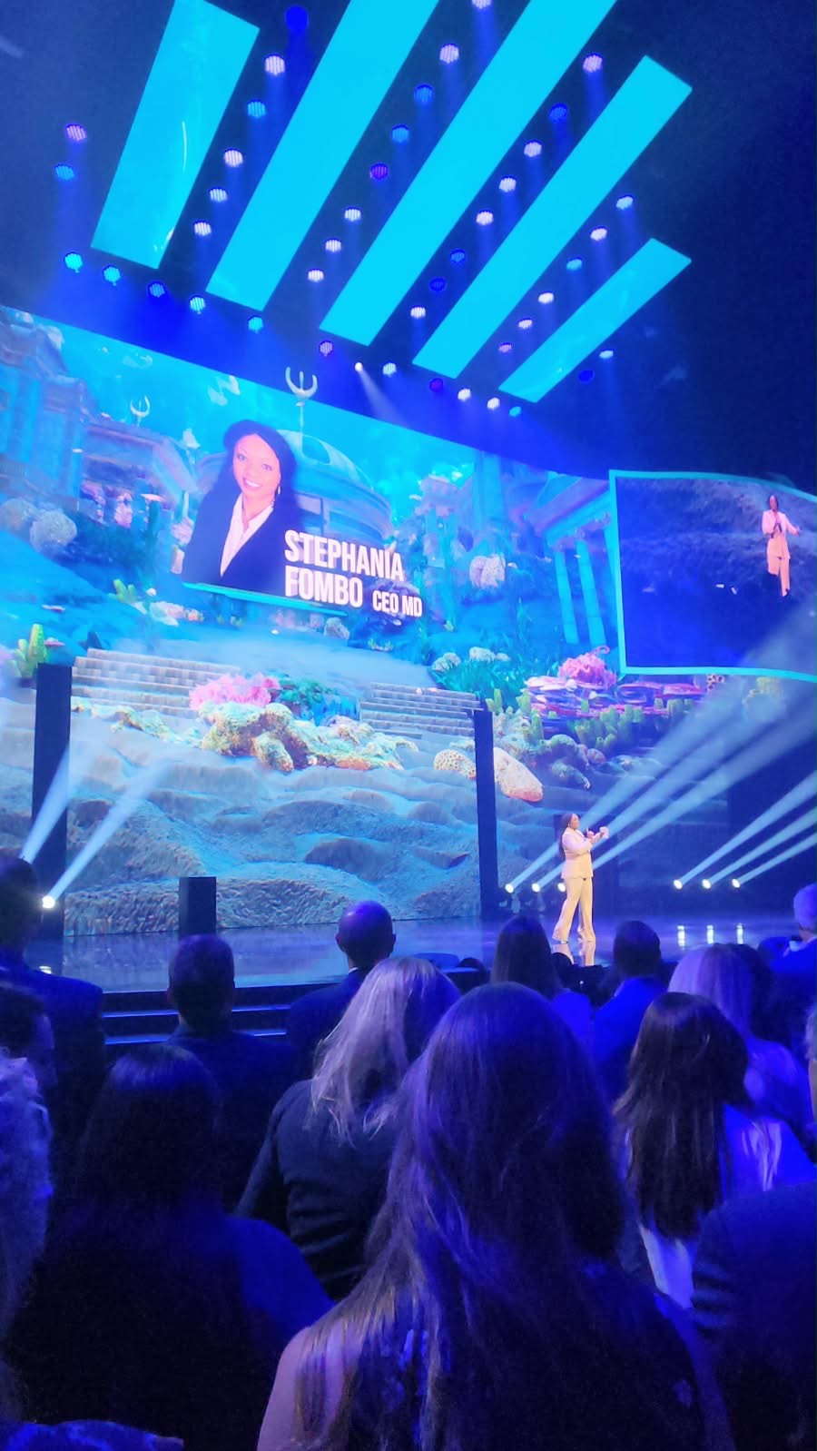 Stephania Fombo speaking on stage at a large event, her name on the screen behind her